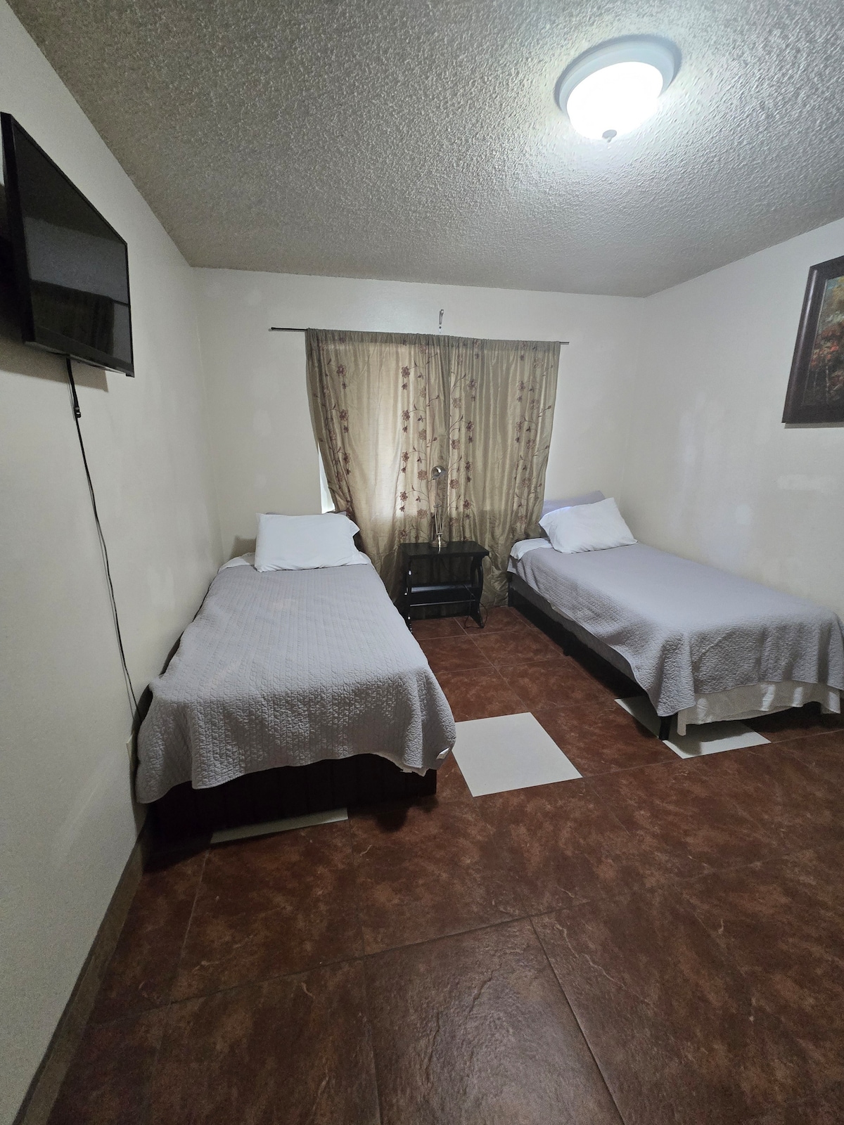 The room features two spacious beds, each dressed in light gray bedding. A television is mounted on the wall opposite the beds, and a dark wooden nightstand is positioned between them. Natural light filters through curtains adorned with a subtle pattern.
