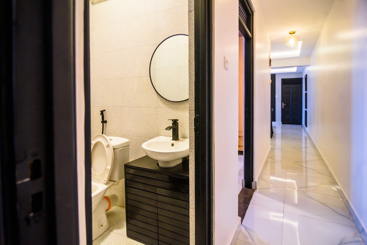 A modern bathroom is visible, featuring a sleek countertop sink with a circular mirror above. A toilet is positioned beside a black storage unit. The area is well-lit with neutral tones and tiled flooring extending into a hallway.