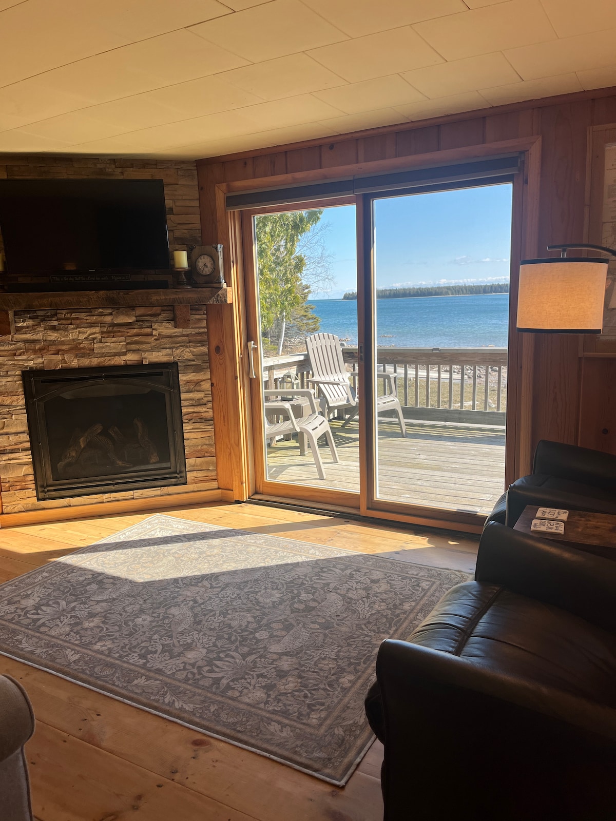 Natural light fills the living area, highlighting a cozy seating arrangement and a stone fireplace. A large sliding door opens to a spacious deck with views of Lake Huron. Comfortable chairs are positioned on the deck, perfect for enjoying the serene waterfront scenery.