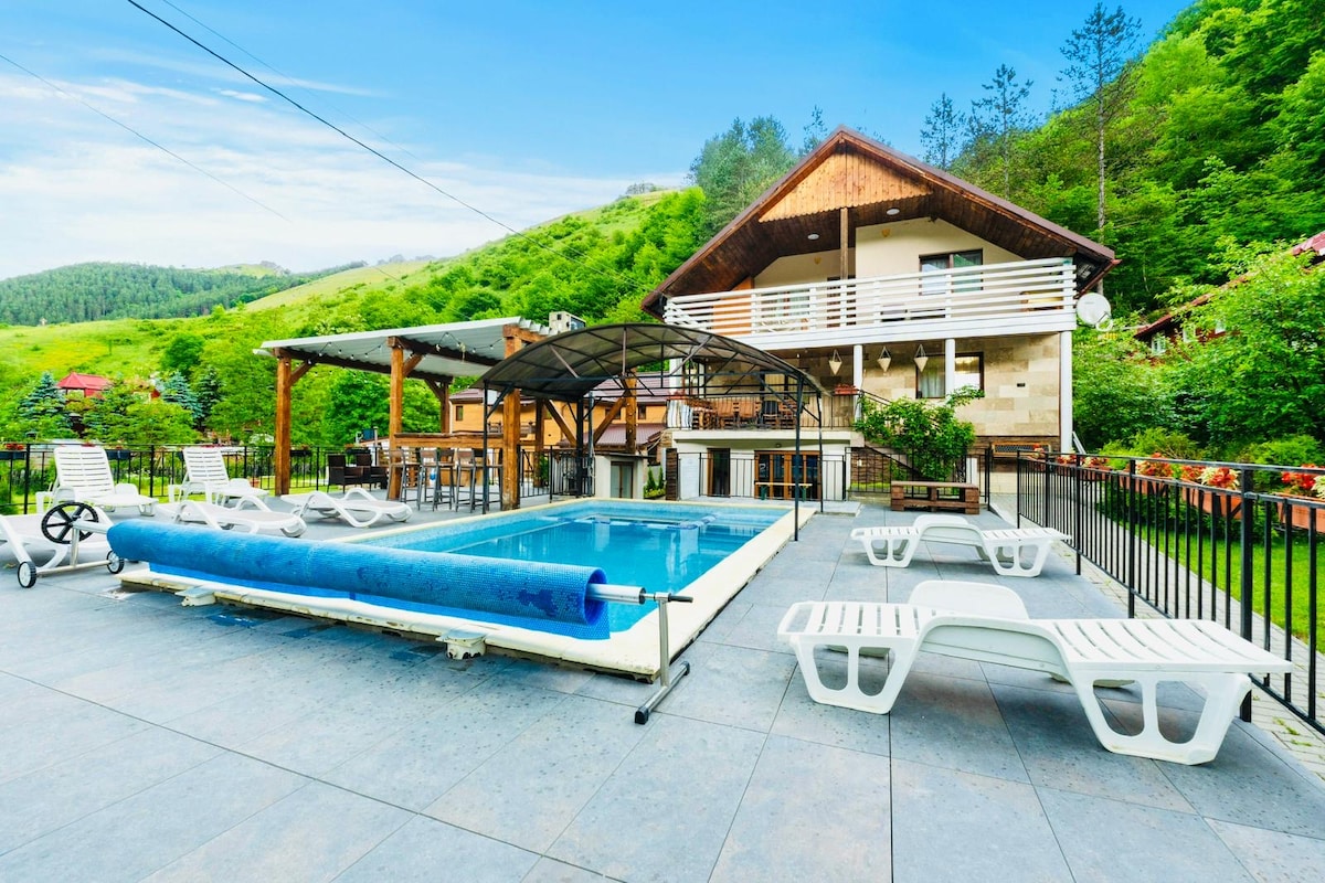 An outdoor area features a swimming pool surrounded by several lounge chairs. A covered seating space with dining options is present nearby, set against the backdrop of green hills. Lush vegetation provides a natural touch, enhancing the outdoor experience.