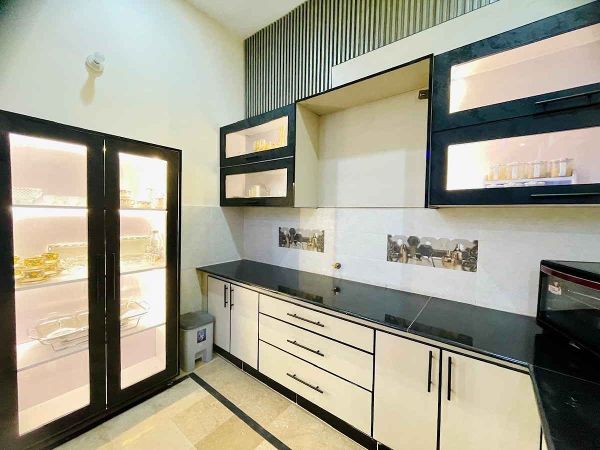 A modern kitchen is showcased, featuring sleek black countertops and cabinetry with contrasting white doors. A glass-fronted cabinet displays various tableware. The space is illuminated by under-cabinet lighting, highlighting the organized layout and stylish design, complemented by a polished stone floor.
