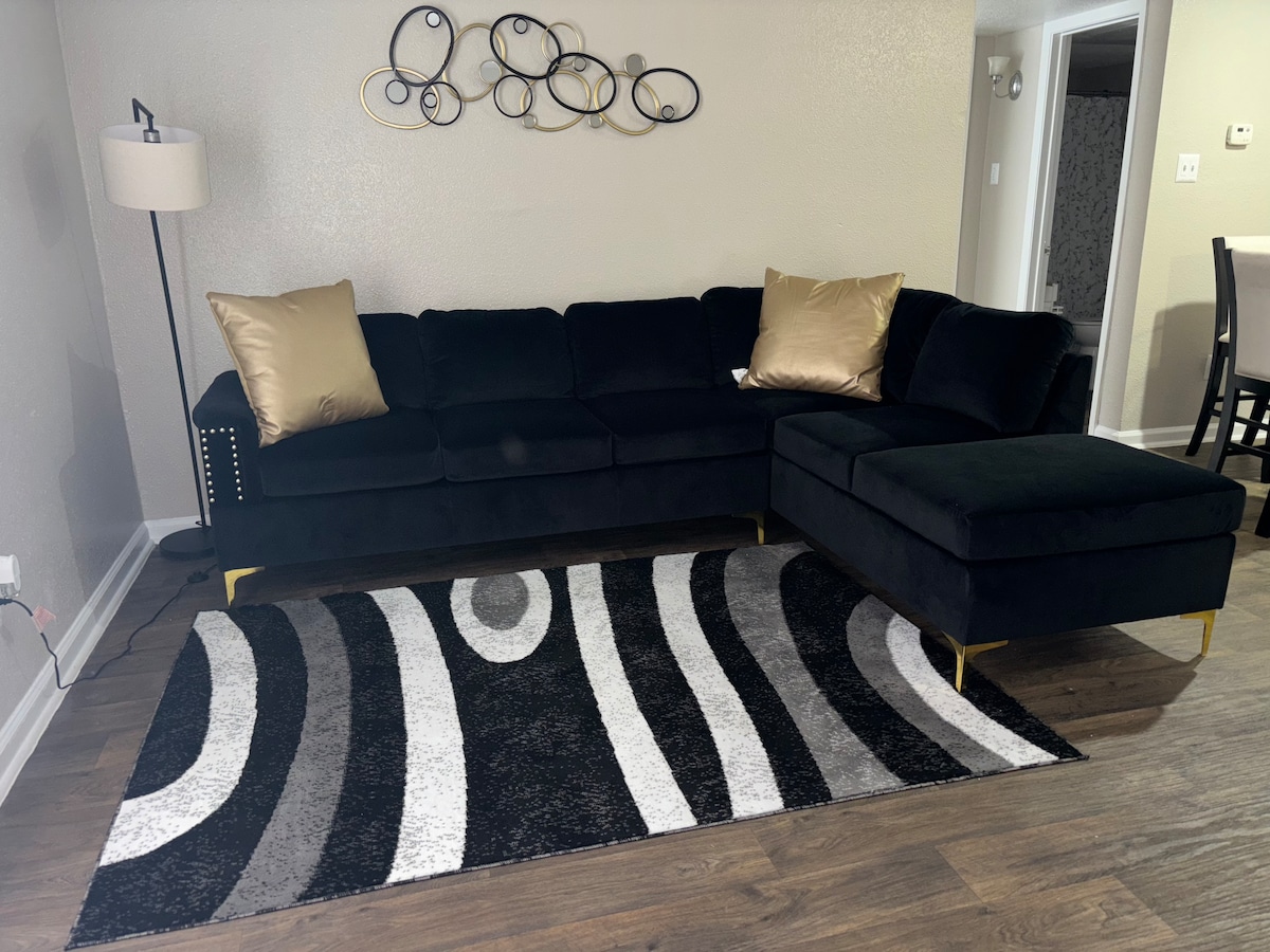 A modern living area features a black sectional sofa, accented with two gold-colored cushions. A patterned area rug with shades of gray, white, and black lies underneath. A lamp provides soft illumination beside the sofa, while a decorative wall piece adds visual interest.