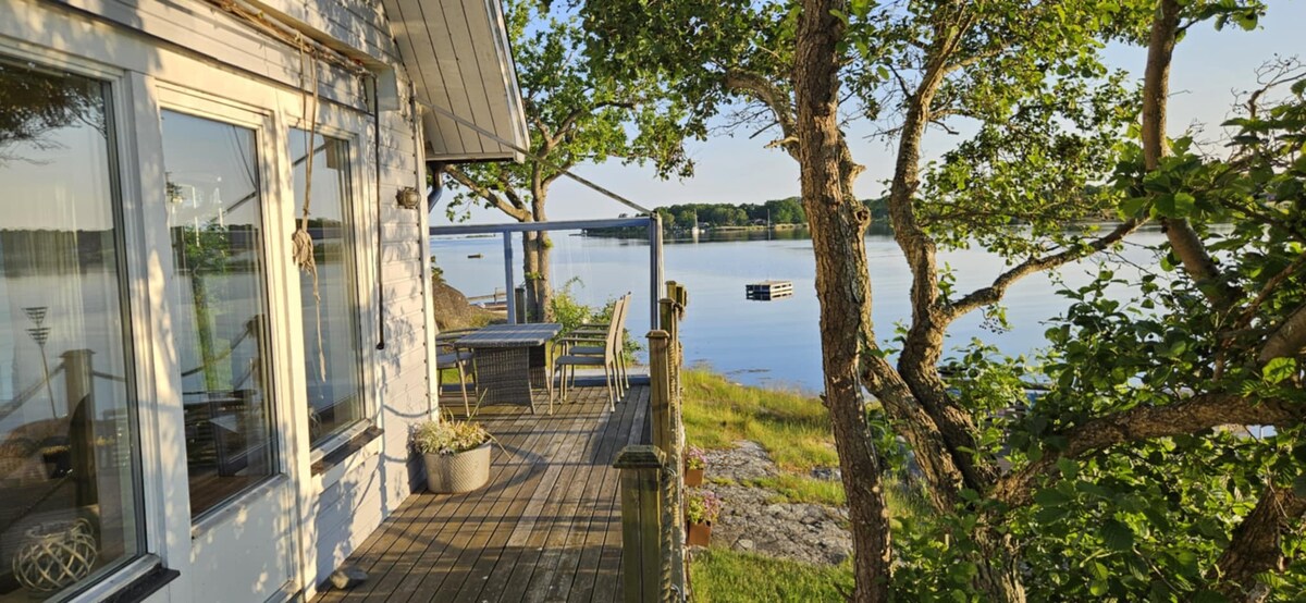 A spacious deck is shown, bordered by trees and overlooking a calm waterway. Seating arrangements and a small table are placed on the wooden surface, inviting relaxation while enjoying the serene view. Sunlight reflects on the water, enhancing the tranquil scene.