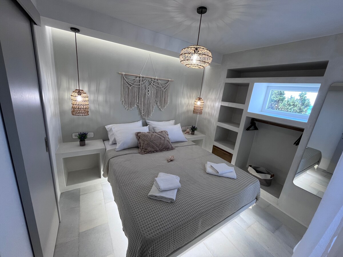 A minimalist bedroom features a spacious bed with neutral-colored bedding. Two woven pendant lights illuminate the room, which includes bedside tables and decorative wall art. A small plant adds a touch of greenery, while the built-in shelves provide storage solutions. Natural light enters through a nearby window.