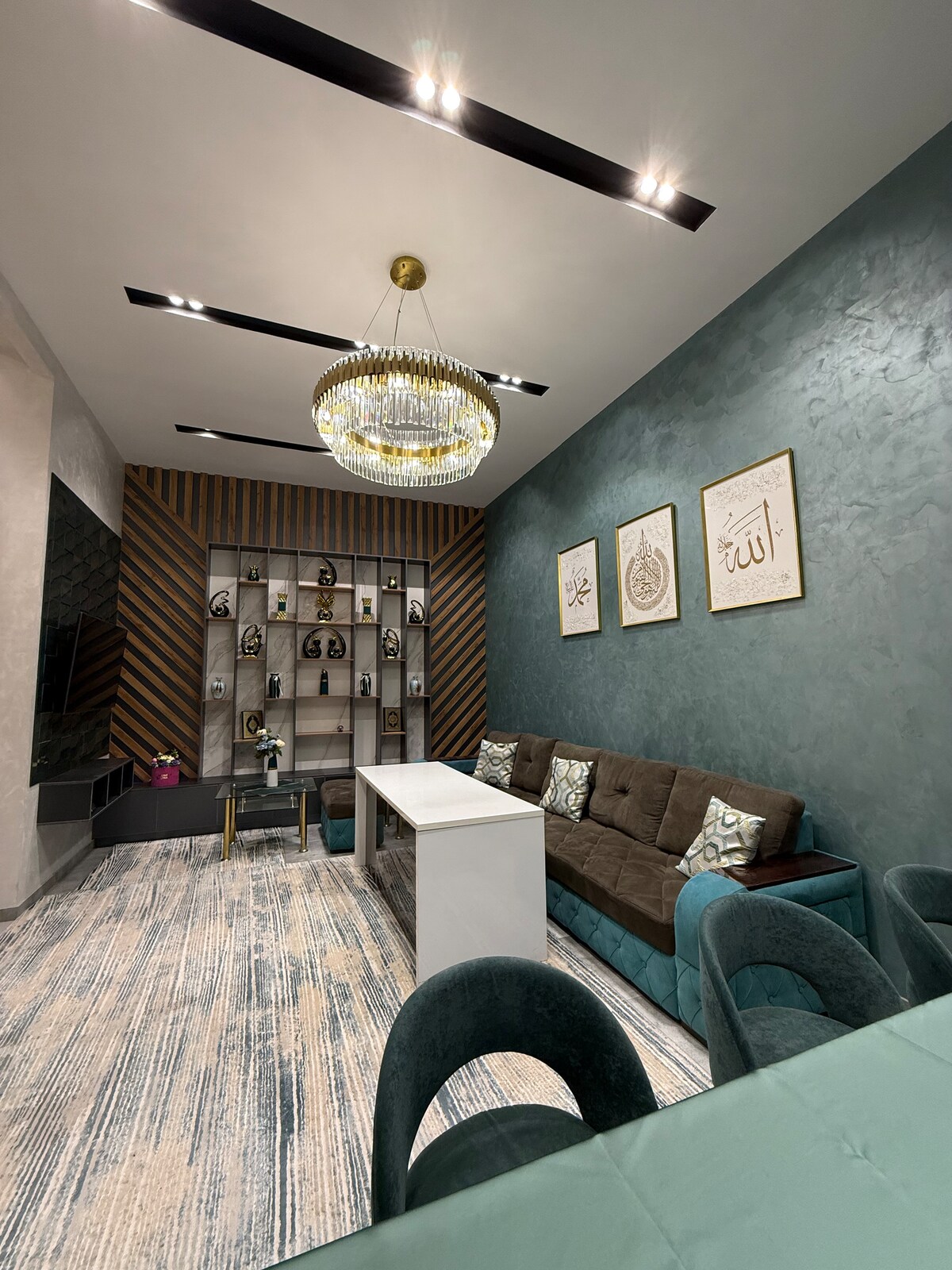 A modern living area features a comfortably arranged seating area with a brown sofa and teal accents. A stylish chandelier illuminates the space, while decorative shelves with various items are displayed against a textured wall. Two framed artworks enhance the overall decor.