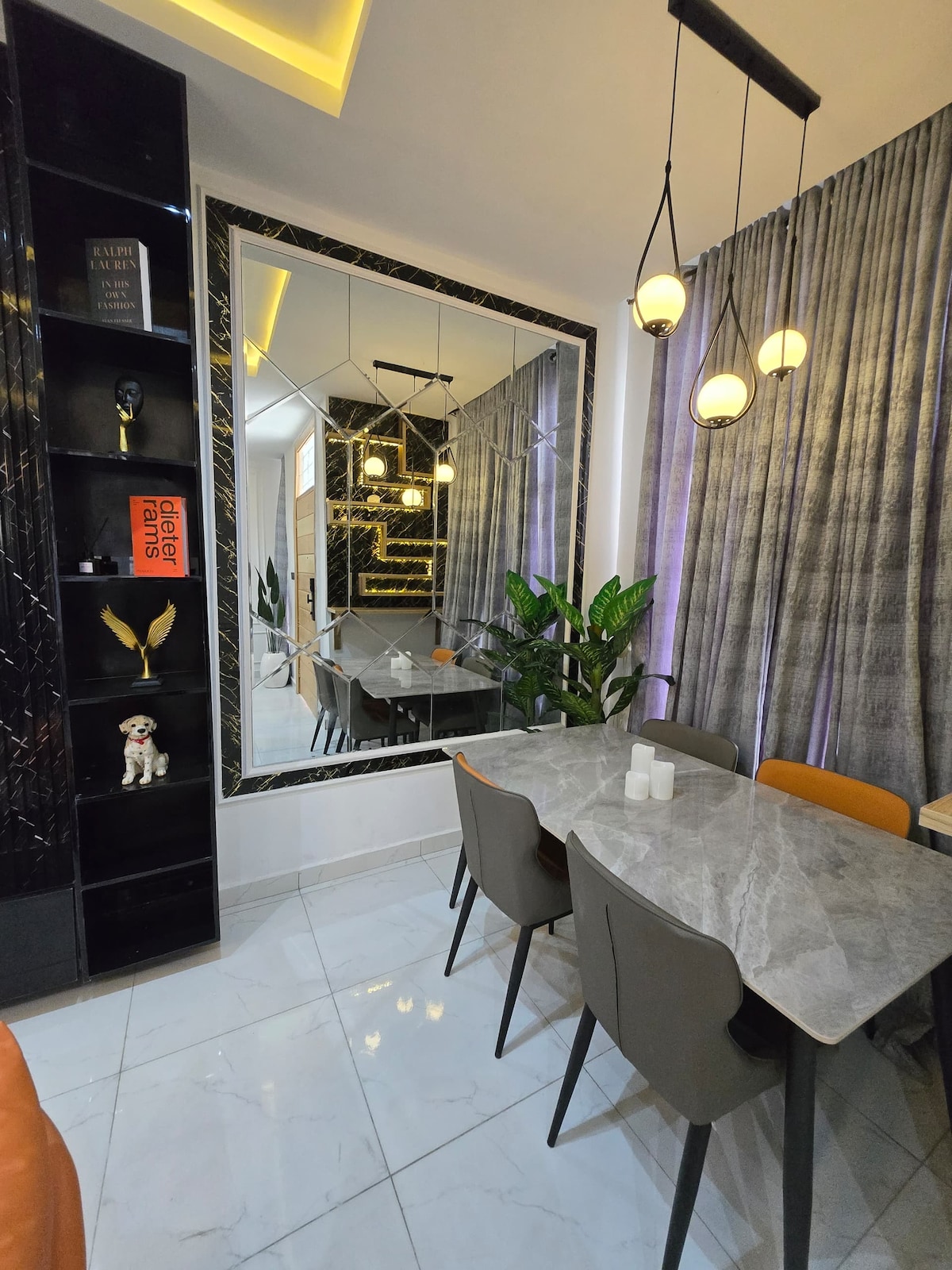 A modern dining area is depicted, featuring a marble table surrounded by sleek chairs. A large mirror amplifies the space's brightness, and green plants add a touch of nature. Elegant lighting fixtures hang from the ceiling, complementing the decor's contemporary style.