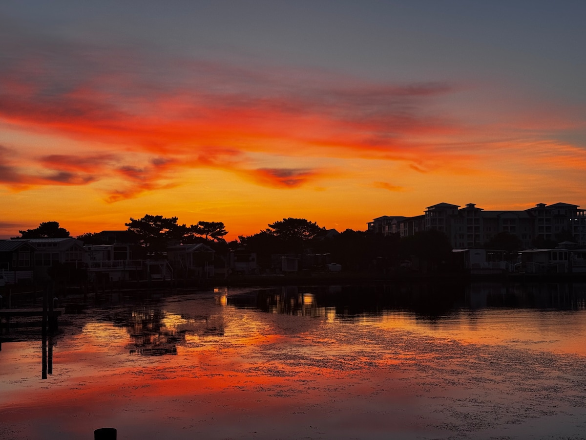 A vibrant sunset paints the sky in shades of orange and pink, reflecting beautifully on the calm water below. Silhouettes of trees and buildings outline the horizon, while the stillness of the scene evokes a serene atmosphere.