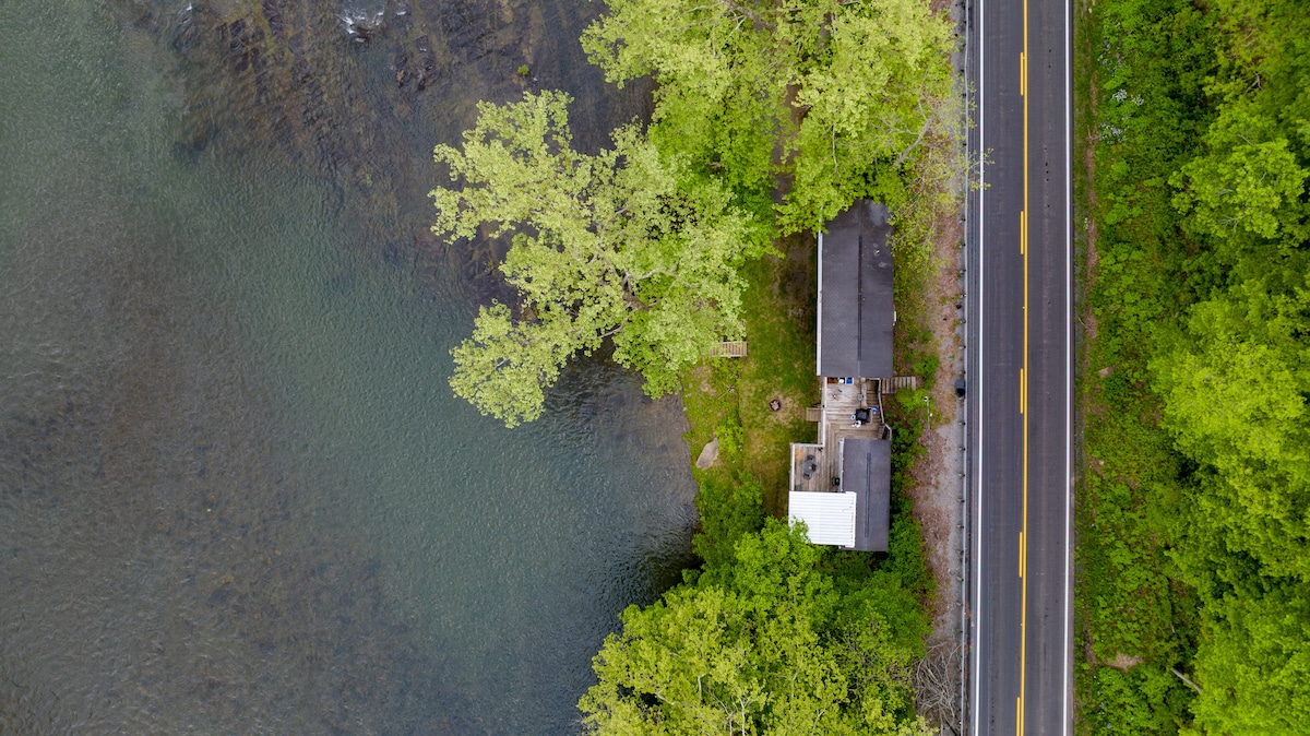 An aerial view captures the cabin nestled beside the Greenbrier River, surrounded by lush greenery. The structure features a simple roof and outdoor seating areas. A road runs adjacent to the river, providing easy access to this tranquil riverside retreat.