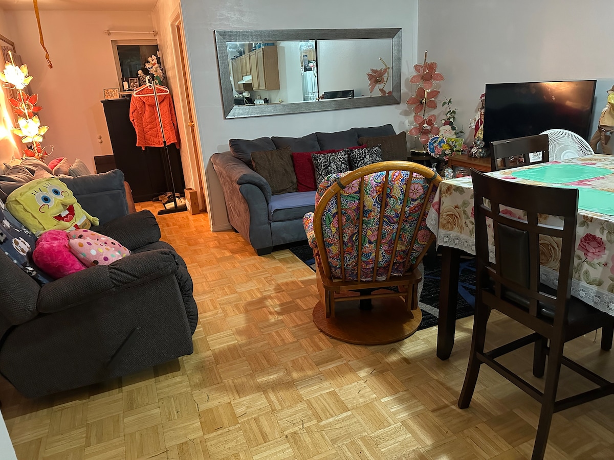 A cozy living area is furnished with a dark gray sofa and a circular rattan chair. A patterned tablecloth covers the dining table surrounded by dark wooden chairs. A large mirror reflects light, enhancing the warm ambiance.