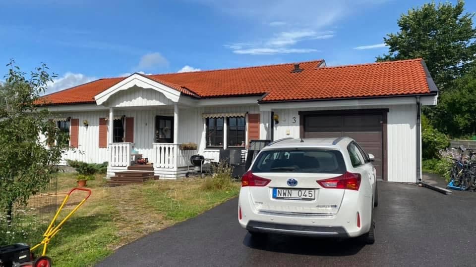 A welcoming house features a red-tiled roof and white exterior with decorative shutters. A driveway leads to an attached garage, while a car is parked in front. A small porch with seating provides an inviting entry. Lush greenery and a bicycle stand are visible nearby.