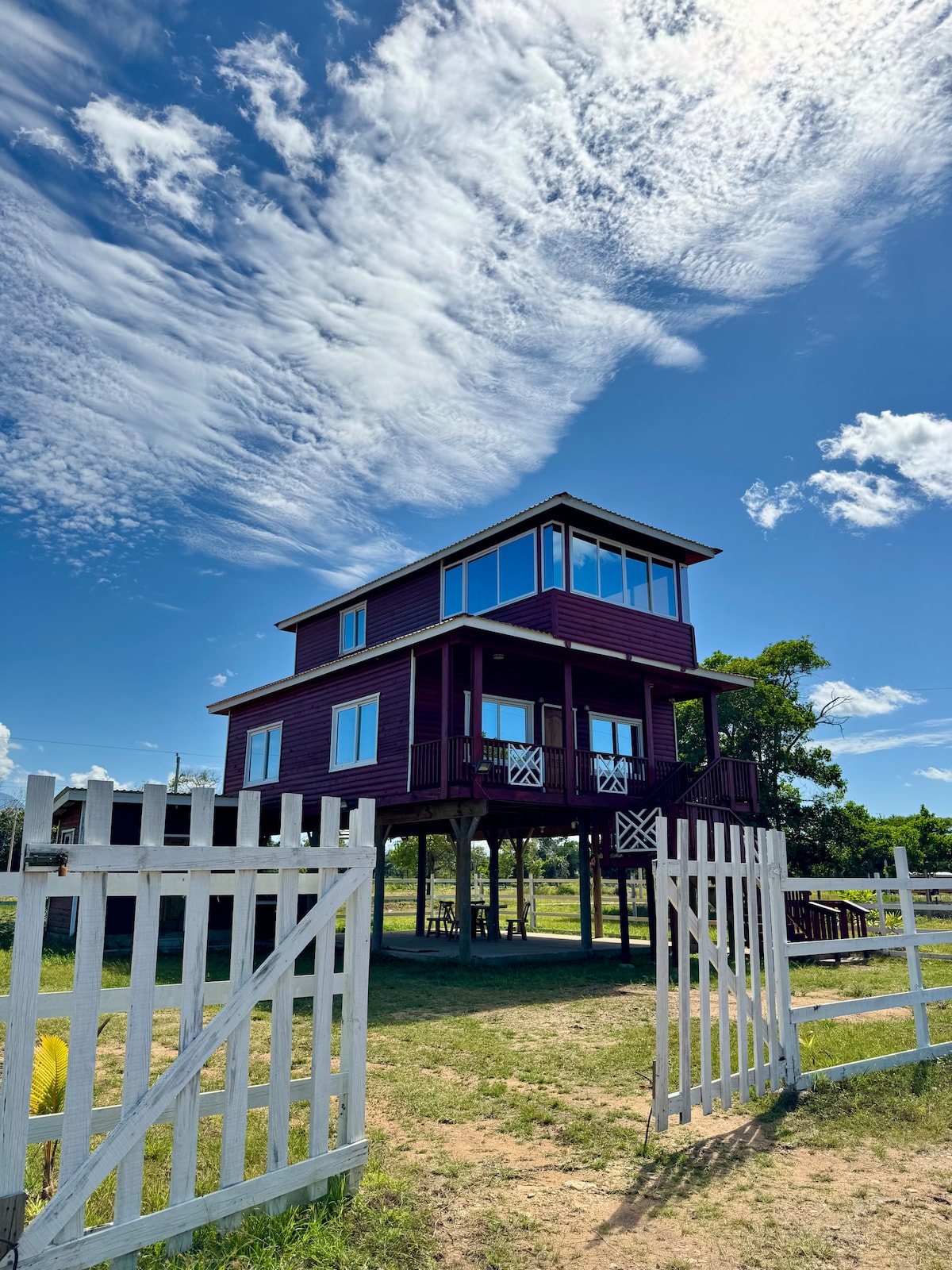A tall, two-story cabin with large windows is seen from behind a white picket fence. The cabin, painted in a deep red hue, is elevated above the ground. The bright blue sky, dotted with wispy clouds, provides a vibrant backdrop.
