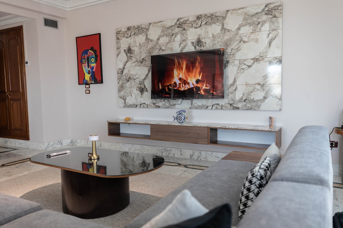 A modern living area is highlighted by a wall-mounted television above a sleek, light-colored console. A stylish coffee table sits in front of a spacious gray sectional sofa, while a vibrant piece of artwork adds color to the neutral-toned decor.