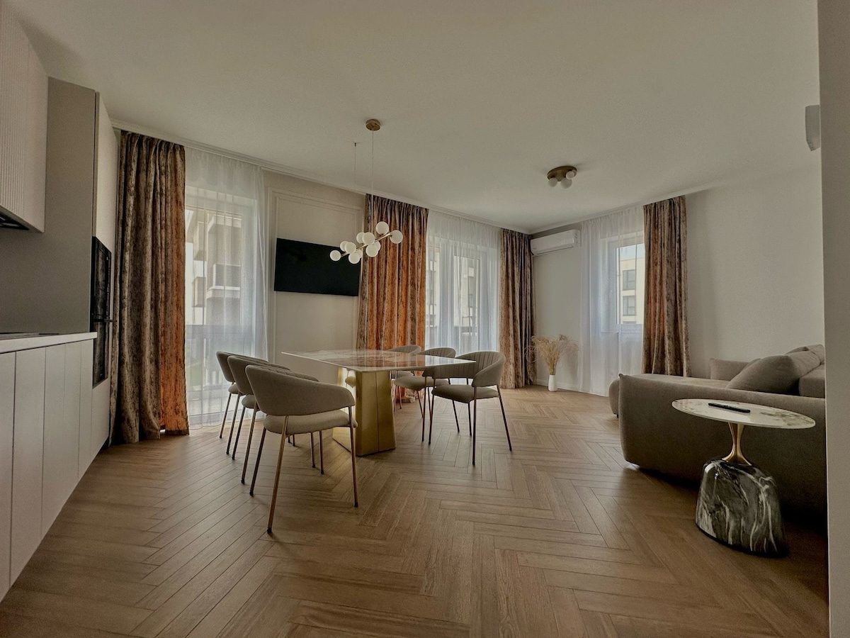 A modern living area features a stylish dining table surrounded by light-colored chairs. Large windows dressed in sheer curtains allow natural light to fill the space. A cozy sofa is positioned in a corner, and a flat-screen TV is mounted on the wall.