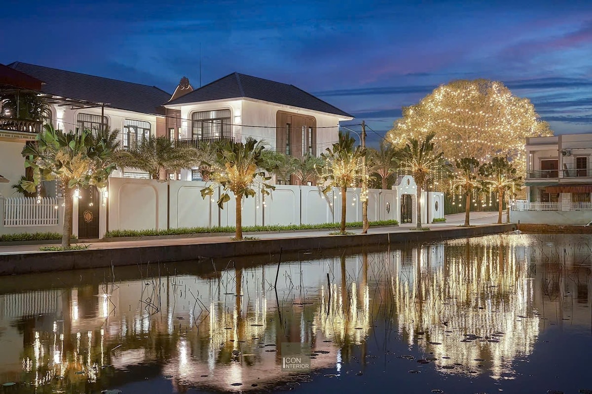 The villa exterior is reflected in the calm water, showcasing palm trees lining the property. Soft lighting adorns the building, highlighting its architectural features against the twilight sky. A large tree in the background is illuminated, adding a gentle glow to the serene scene.