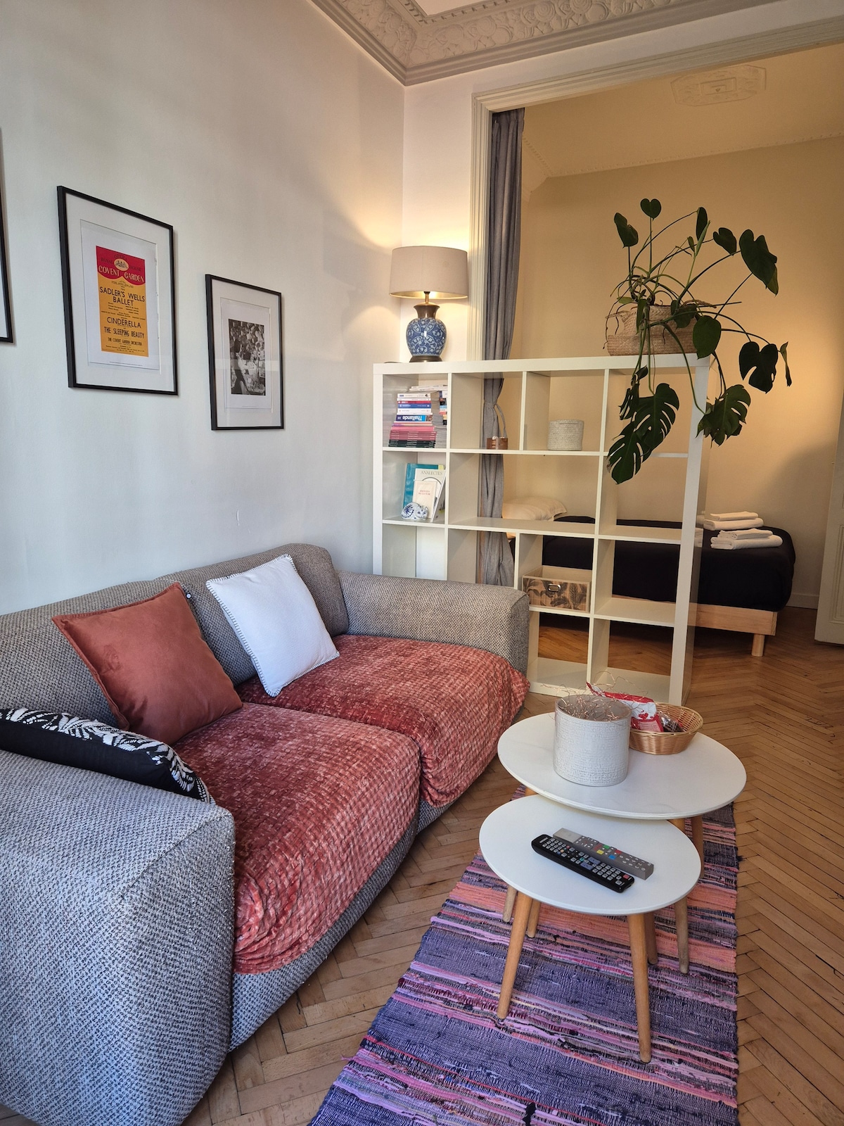 A cozy living area features a textured gray sofa adorned with orange and white cushions. A round coffee table is positioned nearby, accompanied by a small decorative plant and a remote control. Behind, a room is subtly visible, suggesting additional sleeping space.
