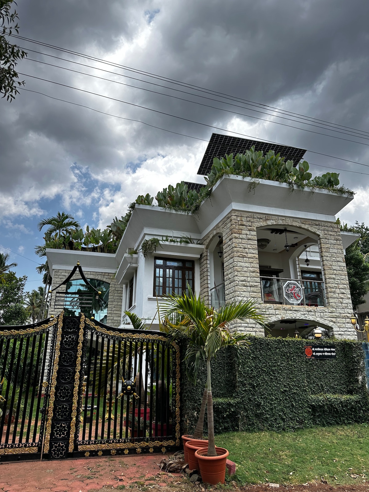 A stylish two-story home features a combination of stone and white walls, adorned with greenery along the rooftop. A gated entrance with ornate black gates opens to a pathway leading to the house, set against a backdrop of cloudy skies and surrounding lush trees.