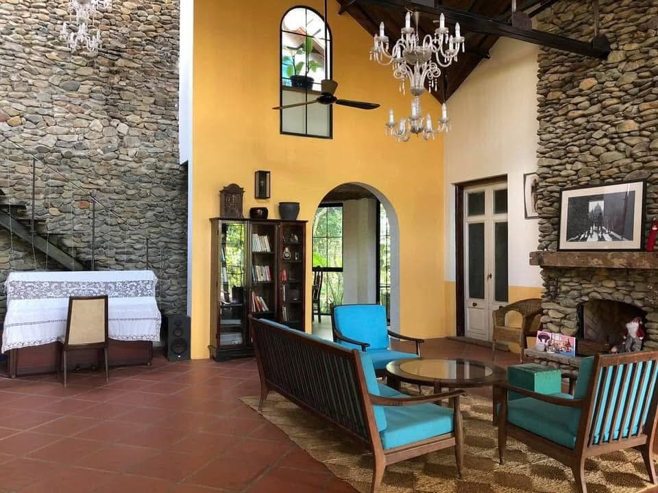 An open living space features a stone accent wall and high ceilings with a wooden beam structure. Comfortable seating is arranged around a central table, complemented by decorative chandeliers. A bookshelf is visible, with greenery seen through an archway leading to the outdoor area.