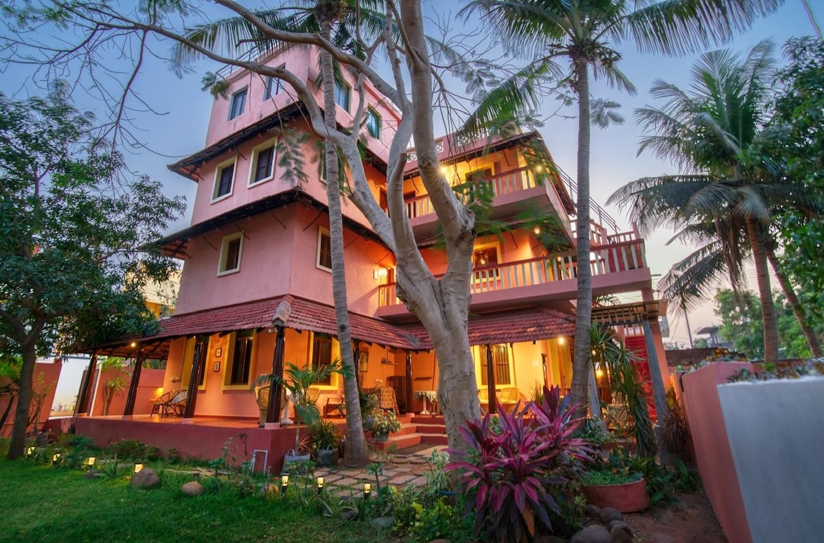 A three-story building is set within a lush garden, featuring vibrant greenery and tropical plants. The exterior is predominantly painted in soft pink tones, with multiple balconies that offer views of the surroundings. Warm light shines through several windows, creating a welcoming ambiance.