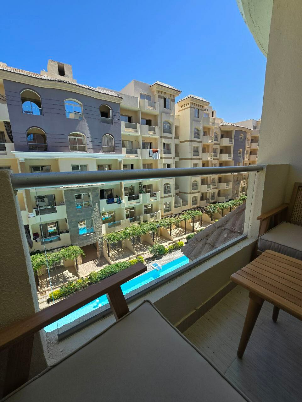 A balcony view reveals a serene outdoor space featuring a swimming pool, framed by well-maintained greenery and residential buildings. Seating is provided by a small wooden table and chair set, positioned to enjoy the sunlit surroundings.