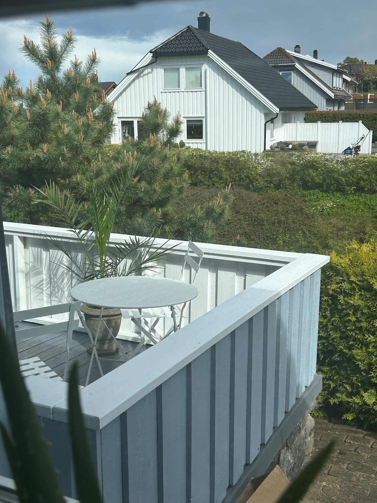 A private balcony is shown, featuring a small round table and two chairs placed against the white railing. Green foliage surrounds the area, providing a tranquil view of the nearby garden and residential houses.