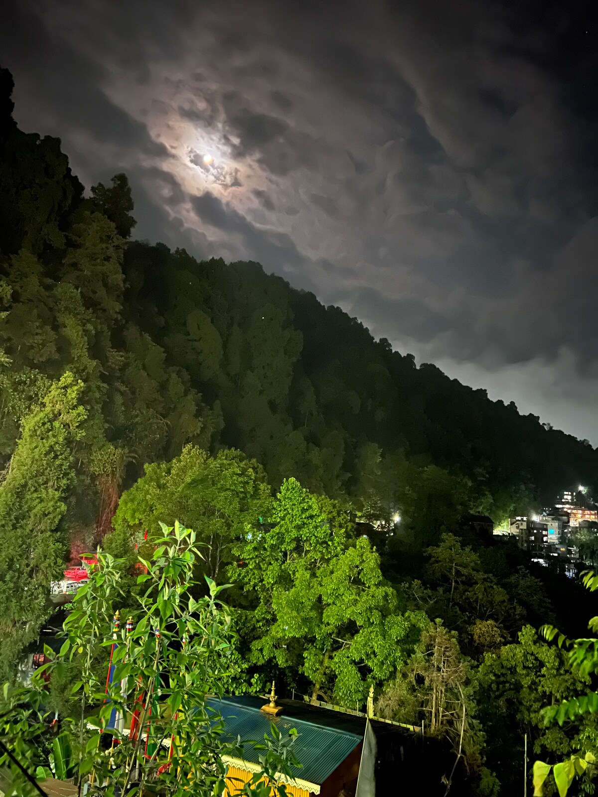 A serene night scene captures a mountainous landscape illuminated by moonlight filtering through clouds. Lush greenery is visible on the hills, while faint lights from nearby buildings add to the tranquil atmosphere, showcasing the peaceful coexistence of nature and civilization.