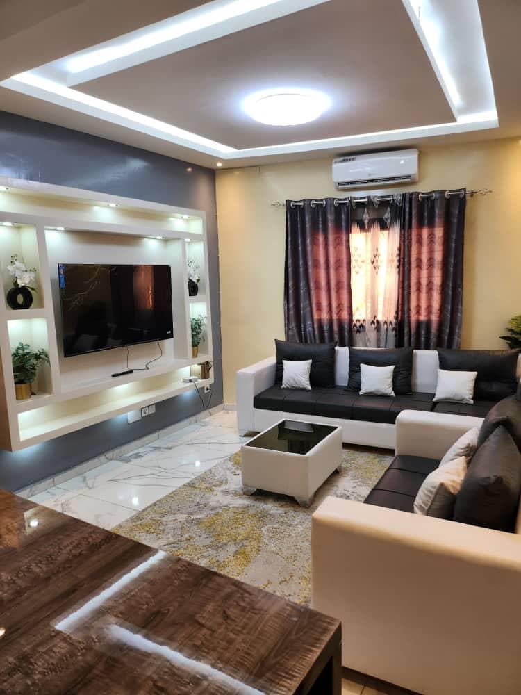 A modern living room is featured, with a large flat-screen television mounted on a backlit wall unit. Comfortable seating includes a white sofa and dark sectional, complemented by decorative pillows. Natural light filters through curtains, highlighting the room's marble flooring and vibrant accent rug.