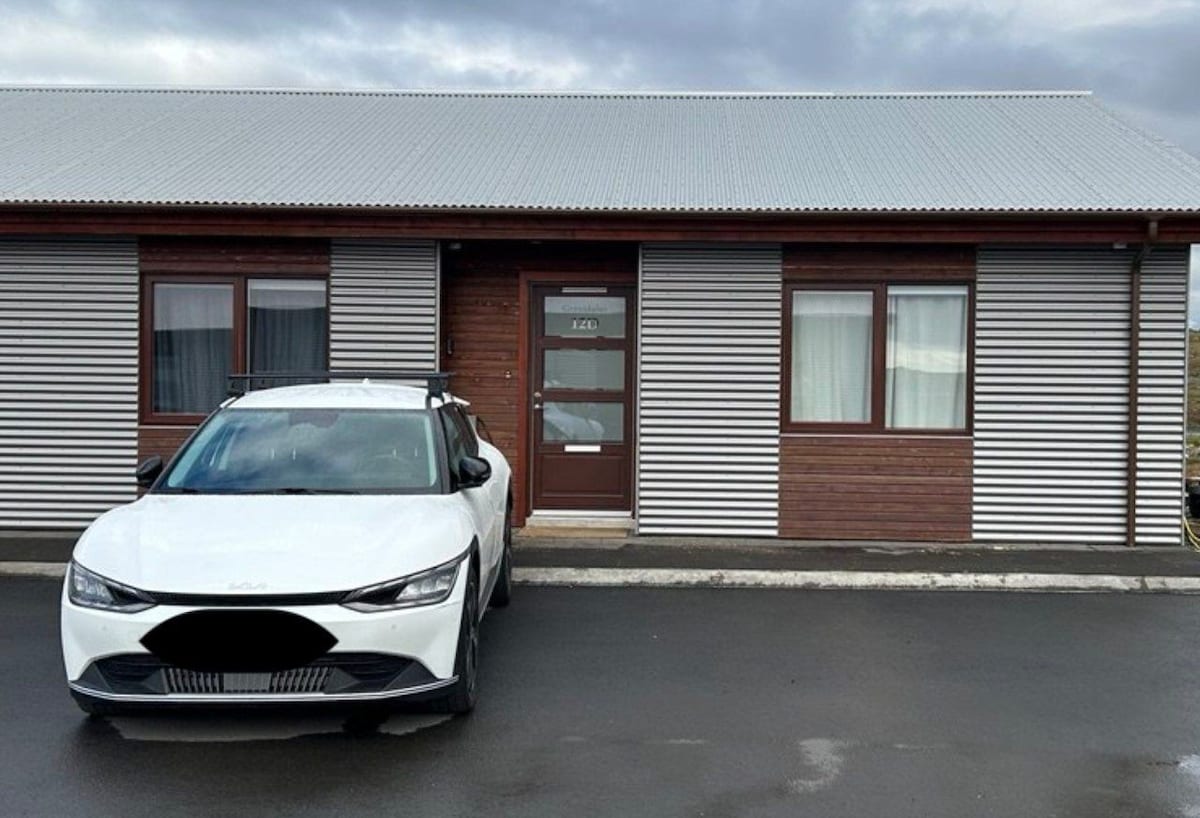 A modern house is shown from the exterior, featuring horizontal wood paneling and a corrugated metal roof. A white vehicle with a roof rack is parked in front, on a paved area leading up to the entrance, which is framed by two windows.