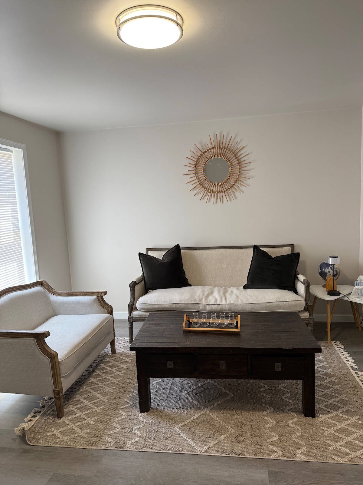 A seating area is showcased with a light-colored sofa and an armchair, both adorned with dark cushions. A dark wooden coffee table sits in the center, accompanied by a decorative tray. A round mirror hangs above the sofa, reflecting natural light from a nearby window.