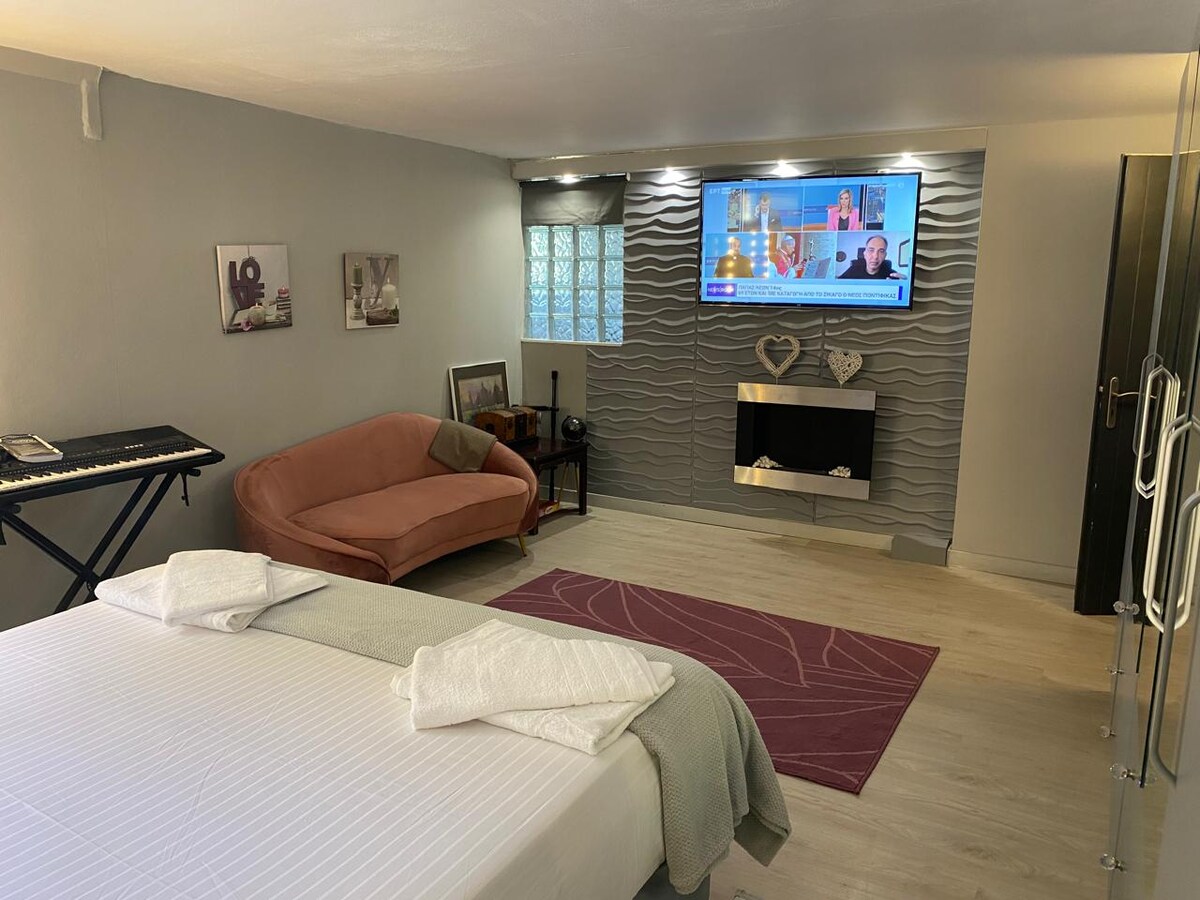 A spacious bedroom is presented, featuring a large bed with neatly folded towels. A soft pink sofa and a keyboard are positioned beside a modern television mounted on a textured wall. Natural light filters through a window with light drapes, creating a calming environment.
