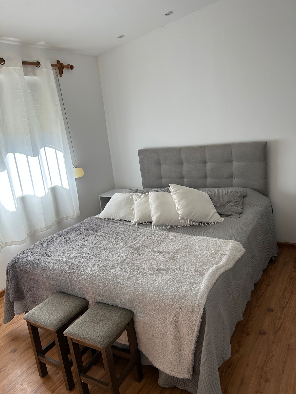 A cozy bedroom is presented with a soft gray bed featuring plush pillows. Natural light enters through a sheer curtain covering a window. Two small stools are positioned at the foot of the bed, adding to the functionality of the space.