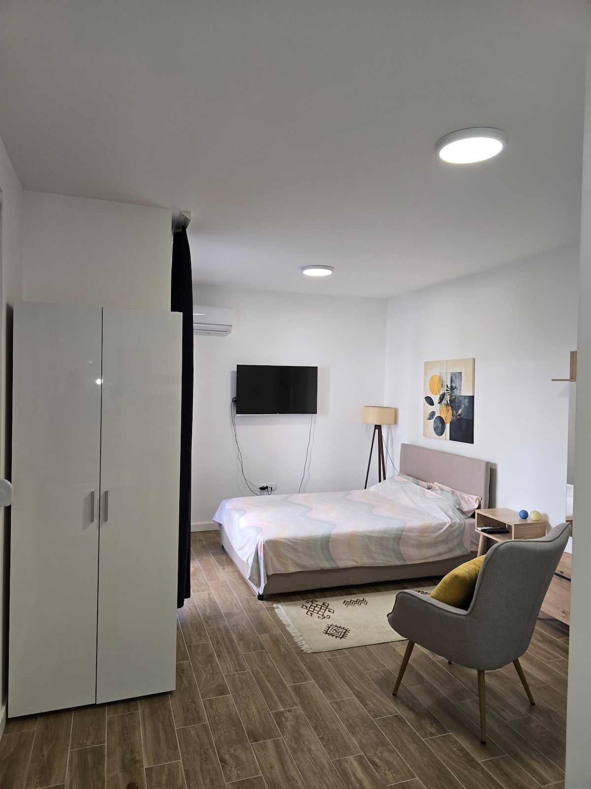 A light-filled sleeping area features a spacious bed with a soft, pale-colored blanket. A flat-screen television is mounted on the wall across from the bed. Accompanying furniture includes a stylish armchair and a tall, white wardrobe. Warm wooden flooring enhances the room's inviting atmosphere.
