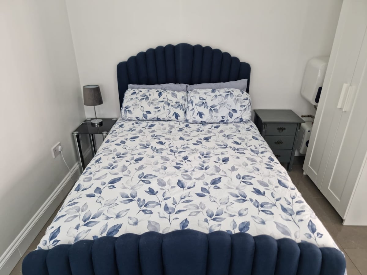 A neatly arranged sleeping area features a spacious bed with a floral-patterned bedding set. Flanking the bed are small nightstands, one with a lamp. A white wardrobe stands against the wall, providing storage options, while soft light brightens the room.