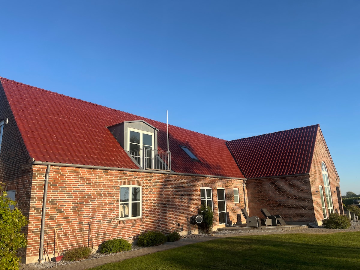 A charming rural building features a red-tiled roof and brick exterior. Large windows allow natural light into the interior. Comfortable seating arrangements are visible outside, surrounded by a well-maintained lawn and garden area.
