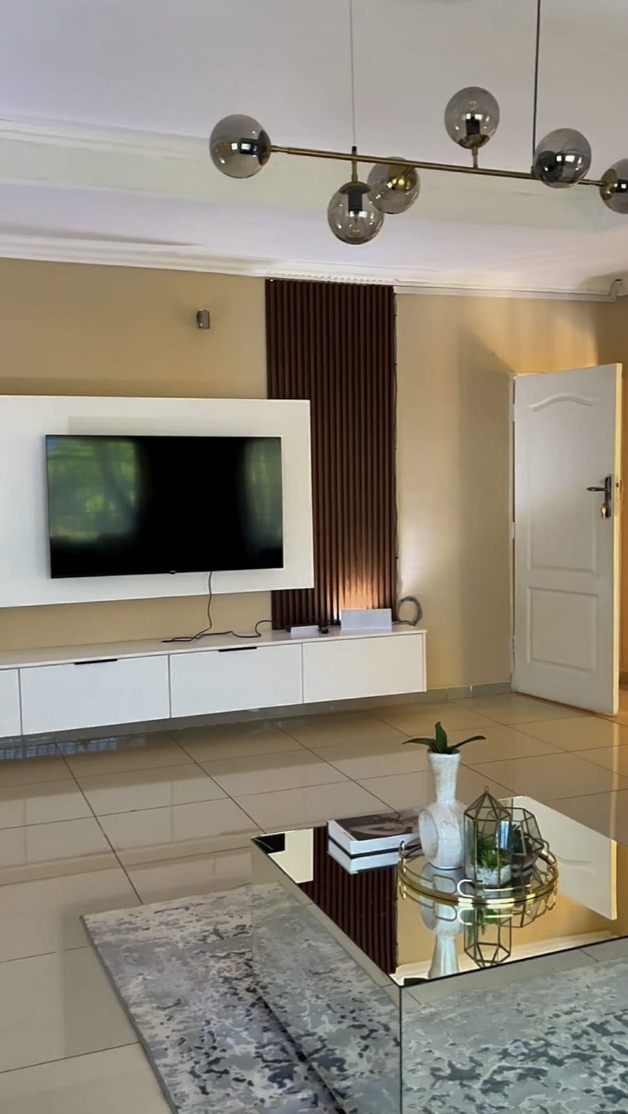 A modern living area is featured with a wall-mounted television above a sleek white cabinet. A reflective glass coffee table occupies the center, surrounded by a light-colored rug. Natural light filters in through a nearby door, illuminating the space.