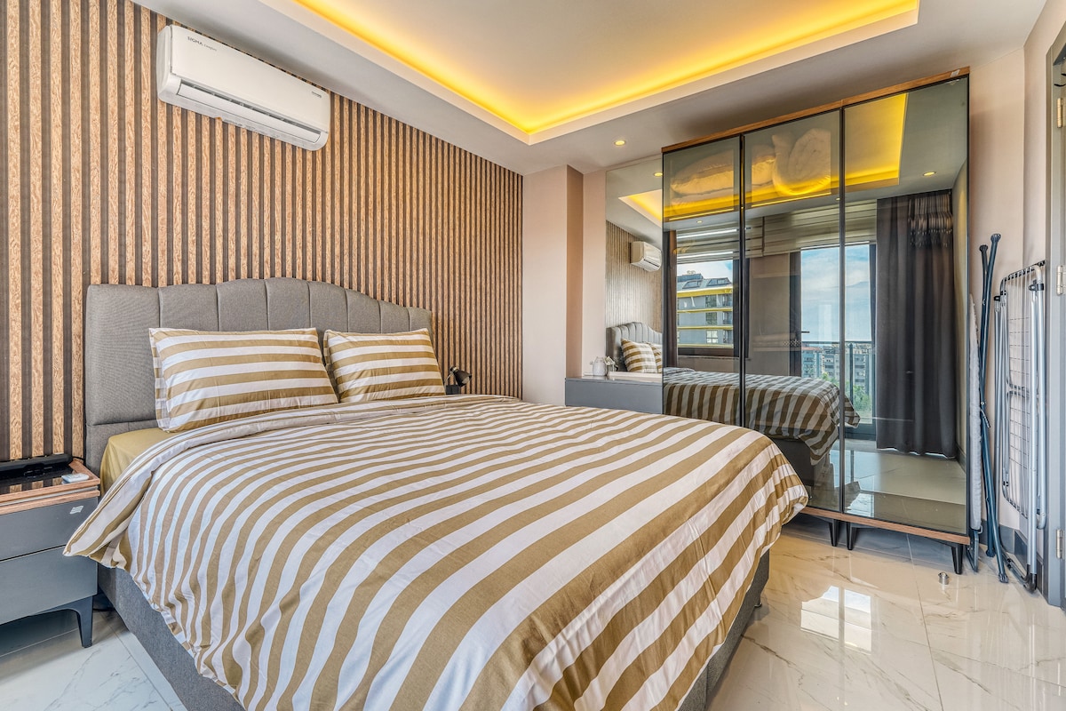 A spacious bedroom is equipped with a large bed dressed in striped bedding. A mirrored wardrobe reflects the light, while large windows provide access to the outside. Soft lighting enhances the calming atmosphere of the room, which features a contemporary design.