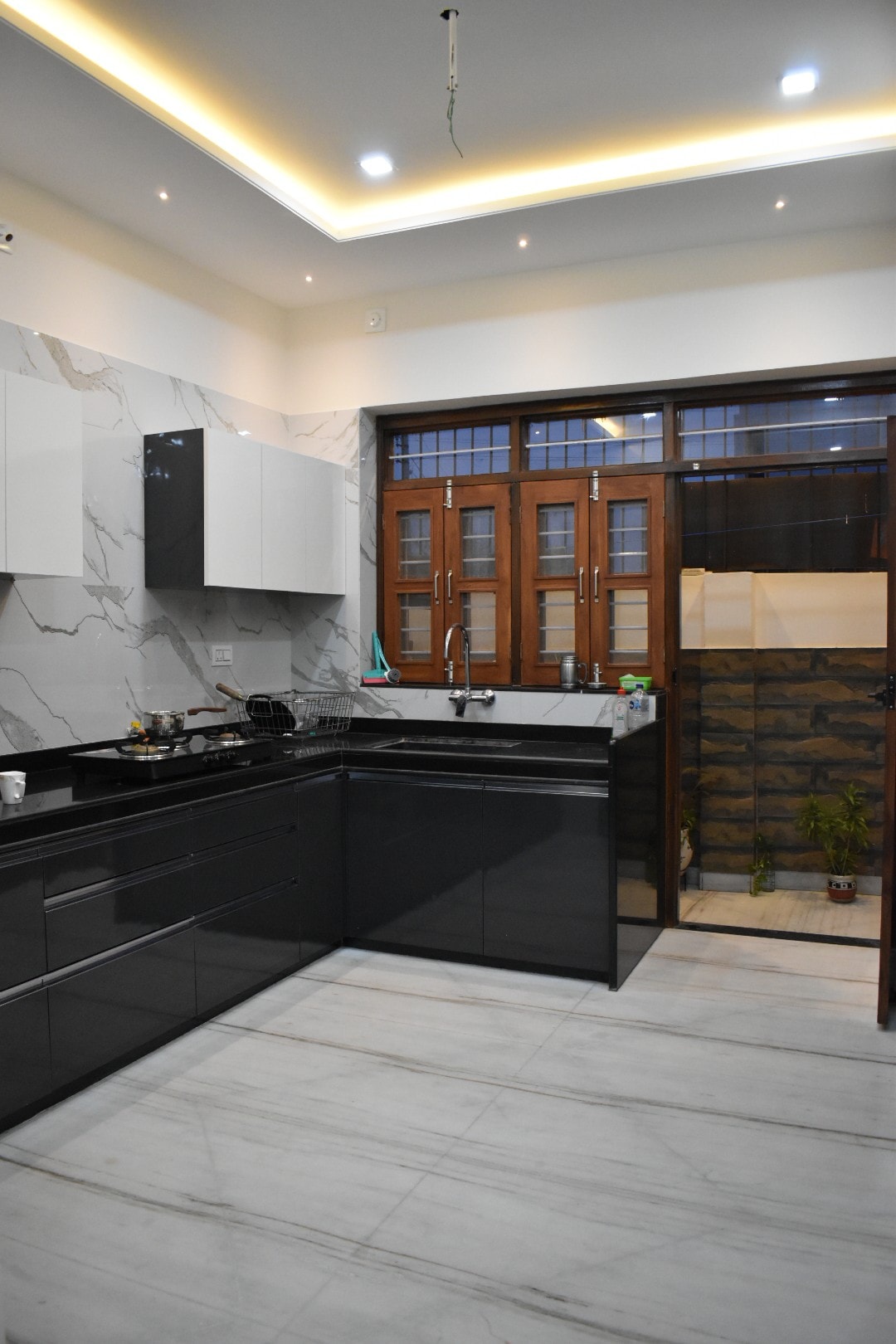 A modern kitchen features sleek black cabinetry and marble countertops, complemented by integrated lighting in the ceiling. Large windows allow natural light to illuminate the space, which includes a small area with plants and a doorway leading outside.