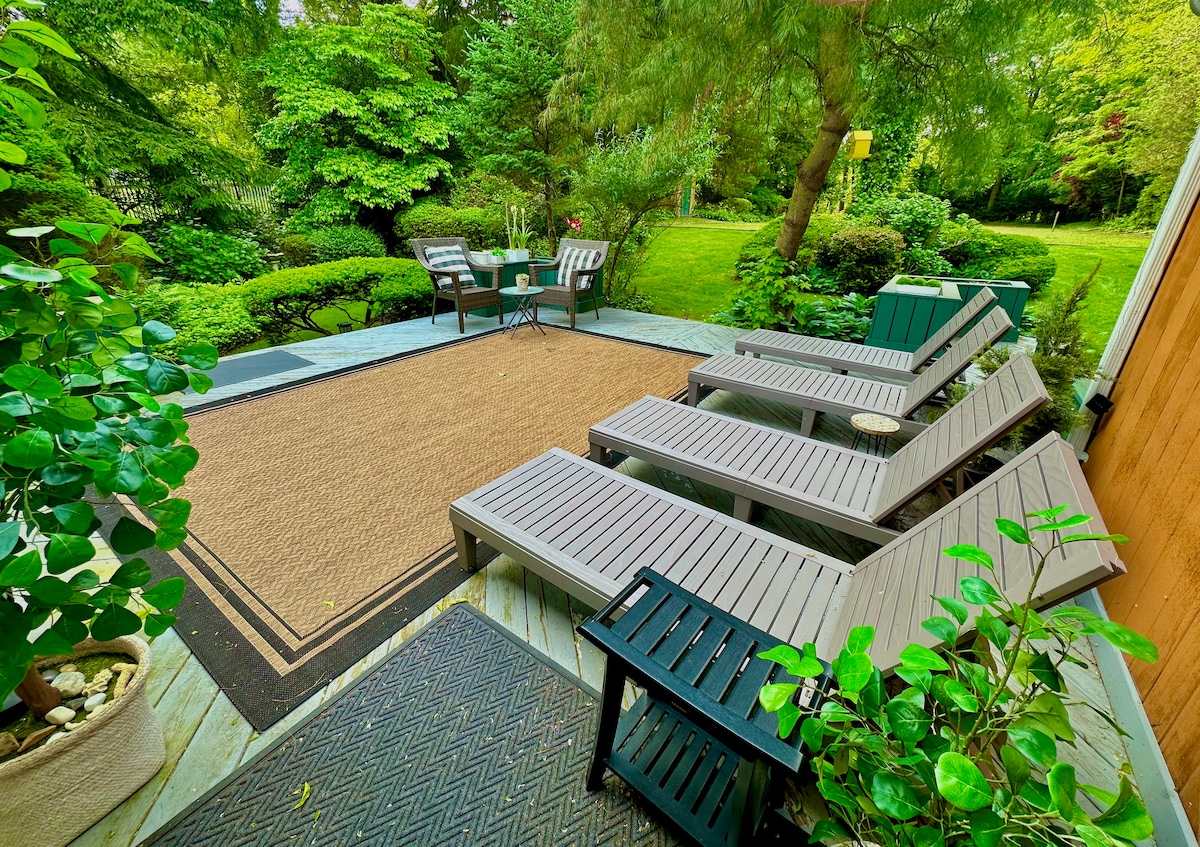 A spacious deck is furnished with multiple chaise lounge chairs arranged around a soft area rug. A small table and chairs are positioned nearby, surrounded by lush greenery and meticulously trimmed bushes, enhancing the tranquil nature of the backyard setting.