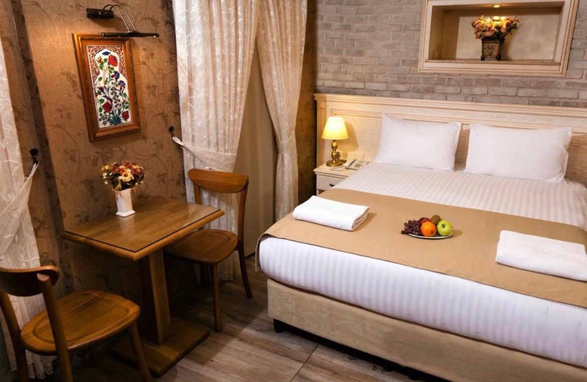 A cozy room features a spacious bed with a neutral-toned bedspread. A small table with two wooden chairs is positioned nearby, accompanied by a decorative vase of flowers. A colorful wall art piece adds character, while soft lighting illuminates the space, creating a welcoming ambiance.