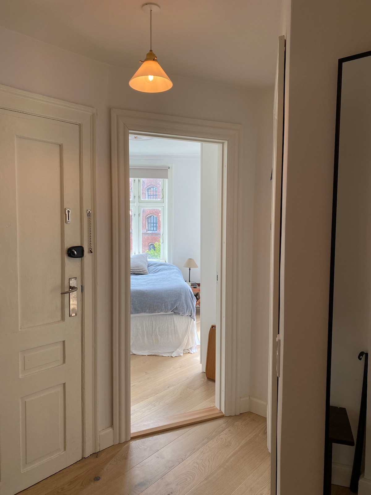 A hallway is presented, featuring a light fixture hanging from the ceiling. A door with a simple white panel is visible, leading to a well-lit bedroom in the background. Natural light brightens the space, highlighting the wooden flooring.