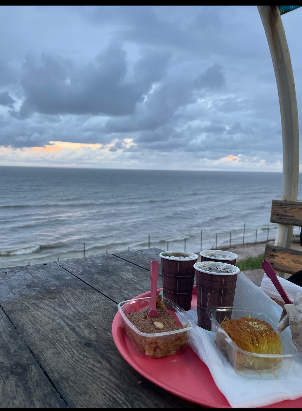 A wooden table is positioned near the shore, displaying three cups of beverage and a plate with assorted desserts. The ocean stretches in the background, with gentle waves under a cloudy sky. Soft colors of the sunset are visible on the horizon.