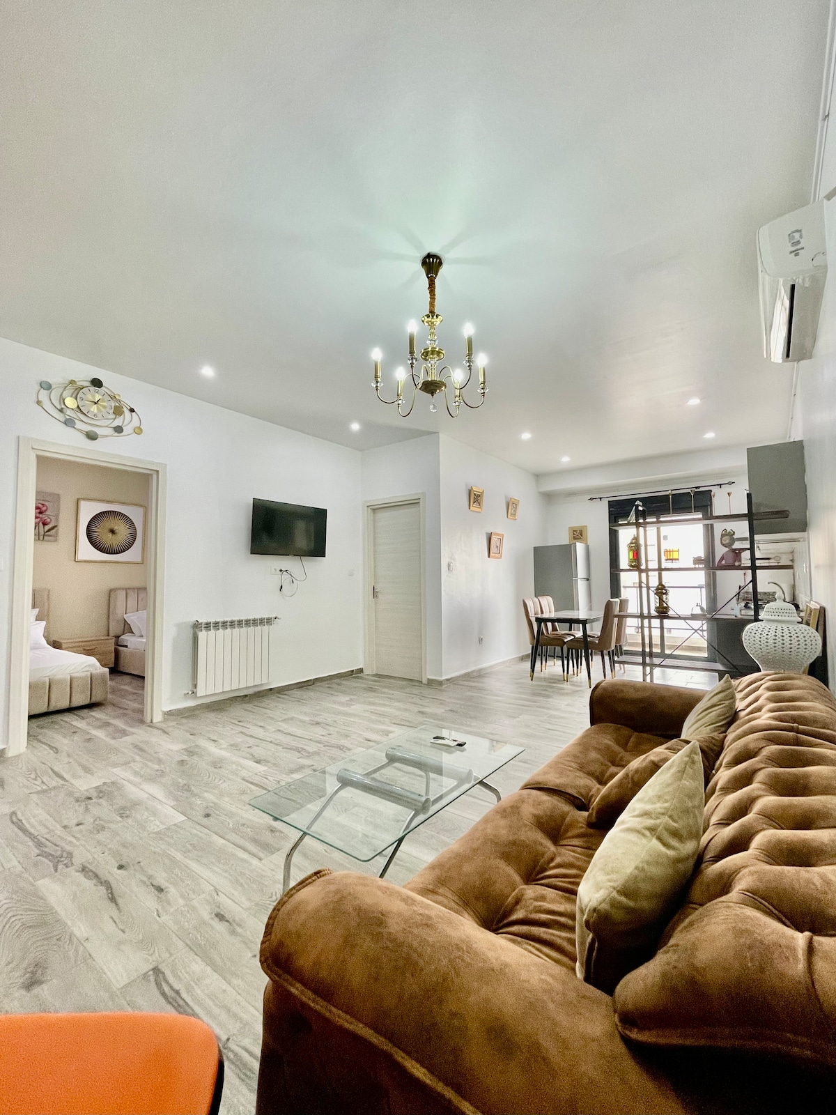 A well-lit living area features a brown velvet sofa, accompanied by a glass coffee table. A wall-mounted TV is visible, and a dining space with wooden chairs is positioned nearby. Bright light streams in from a large window, enhancing the open layout.