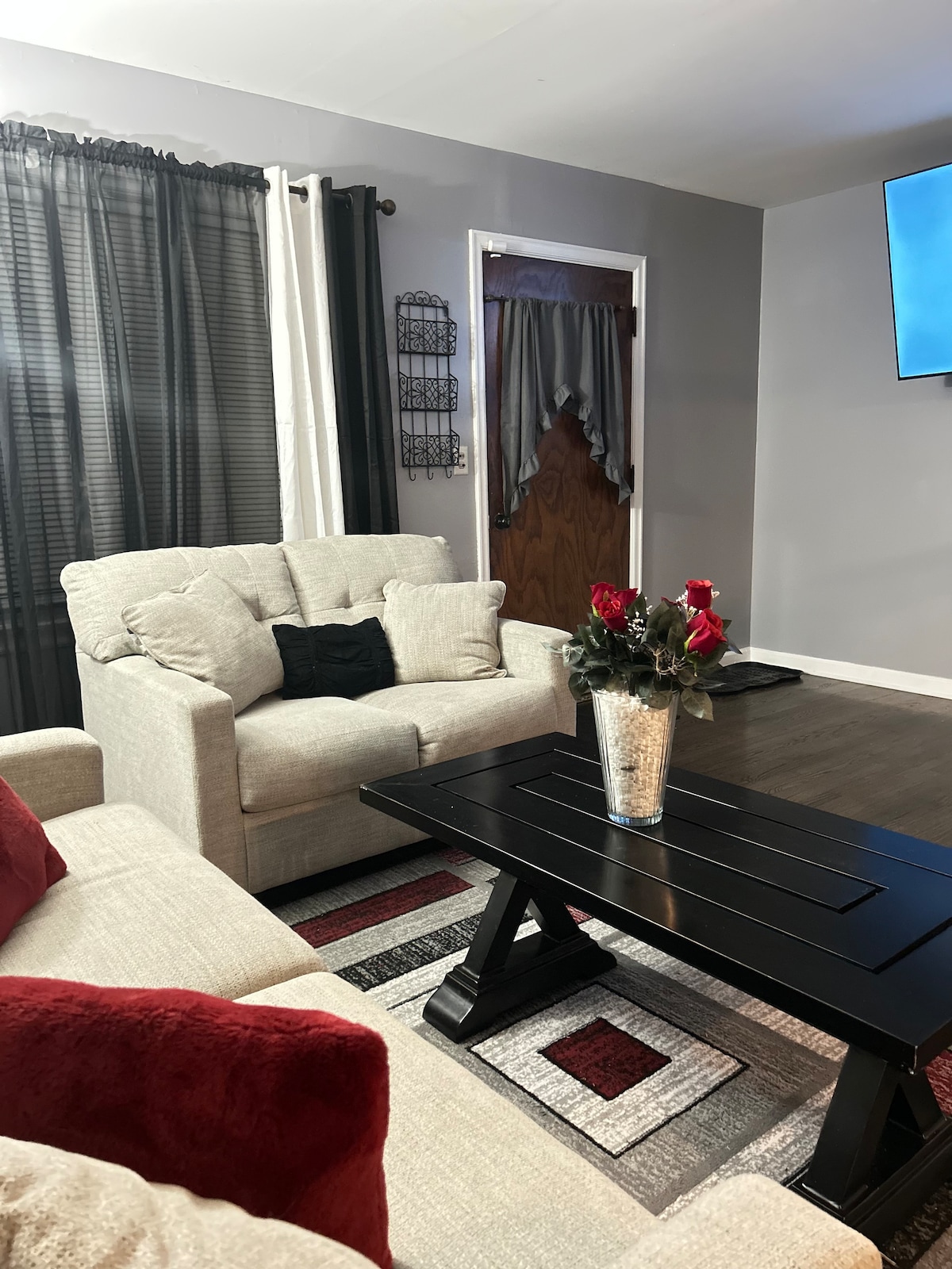 A comfortable living area is presented, featuring a light-colored sofa and armchair positioned around a dark wooden coffee table. A vase with flowers decorates the table. Soft lighting from a wall-mounted television adds a relaxed feel to the space. Dark curtains frame the window.
