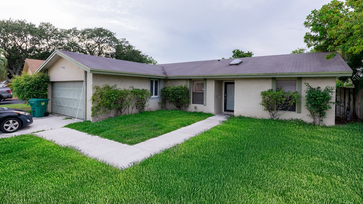 The exterior of the home is shown, featuring a well-maintained yard with lush green grass. A concrete path leads to the front entrance, flanked by small shrubs. The single-story structure includes a garage and has a simple yet inviting façade.