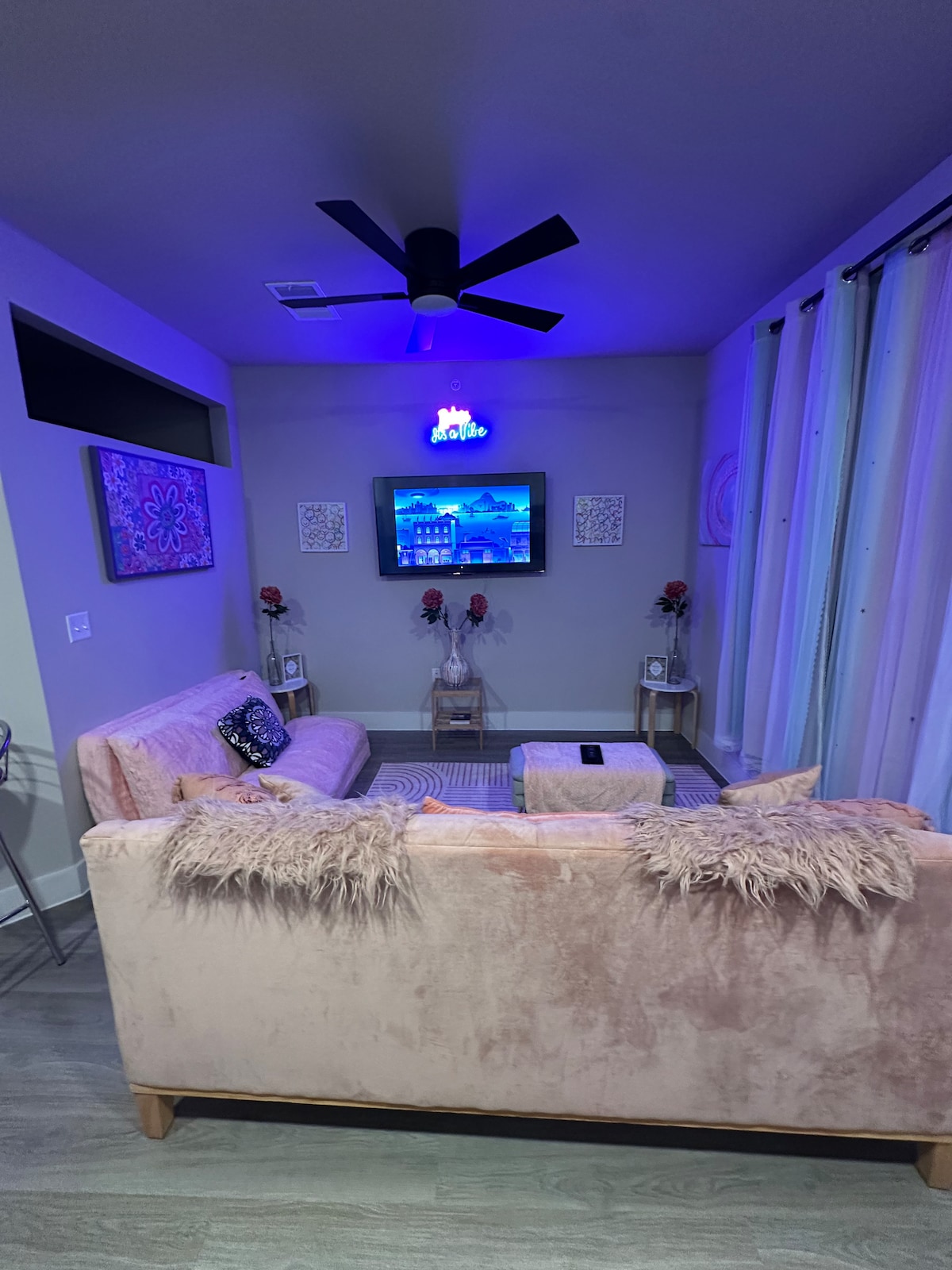 A softly lit living area features a light pink sofa adorned with textured throws. A low table is positioned in front, while a ceiling fan contributes to airflow. Decorative elements include wall art and lamps alongside the seating, with a television mounted on the wall displaying scenery.