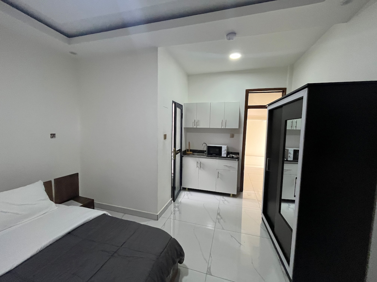 The spacious studio features a queen bed with neutral bedding and wooden headboard. A kitchenette with essential appliances is visible, including a microwave and storage cabinets. A large cabinet and a modern light fixture complement the minimalist design, while a bright ambiance is created by overhead lighting.