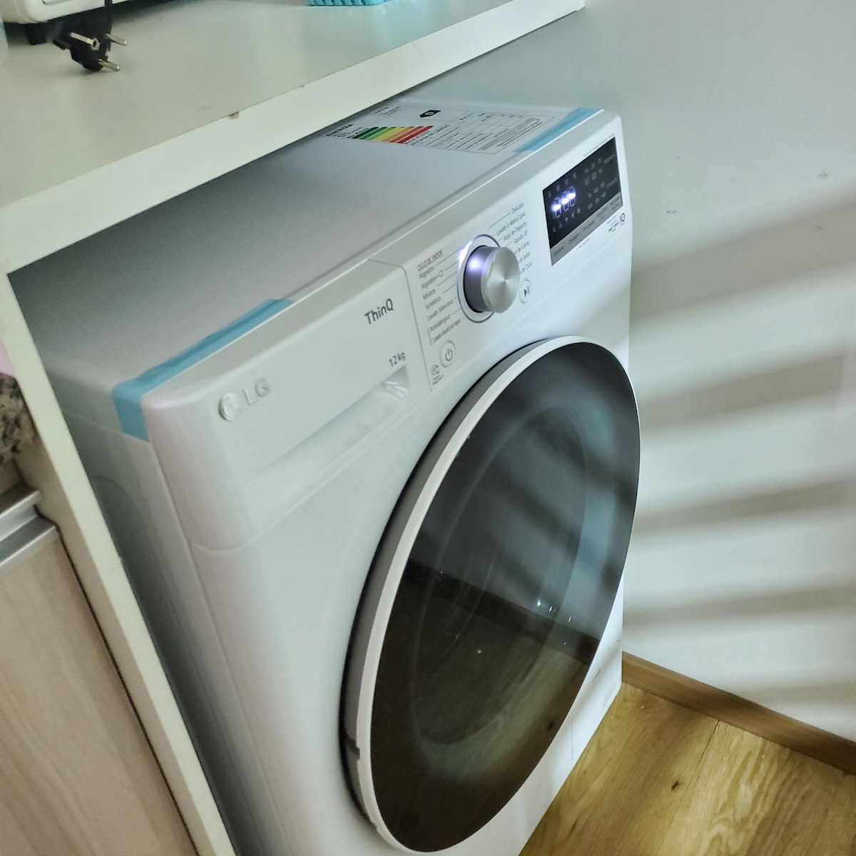 A modern washing machine is positioned under a countertop, featuring a sleek white design. Control buttons and a digital display are located on the front, while the door is partially transparent, allowing a view of the interior. Clean wooden flooring complements the space.