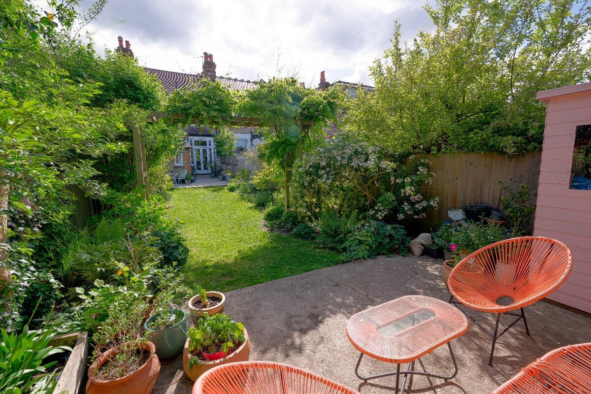 A private garden is featured, showcasing lush greenery and flowering plants. Terracotta lounge chairs surround a glass-topped table on the patio. The garden leads to a grassy area, framed by trees and shrubs, offering a peaceful setting for relaxation.