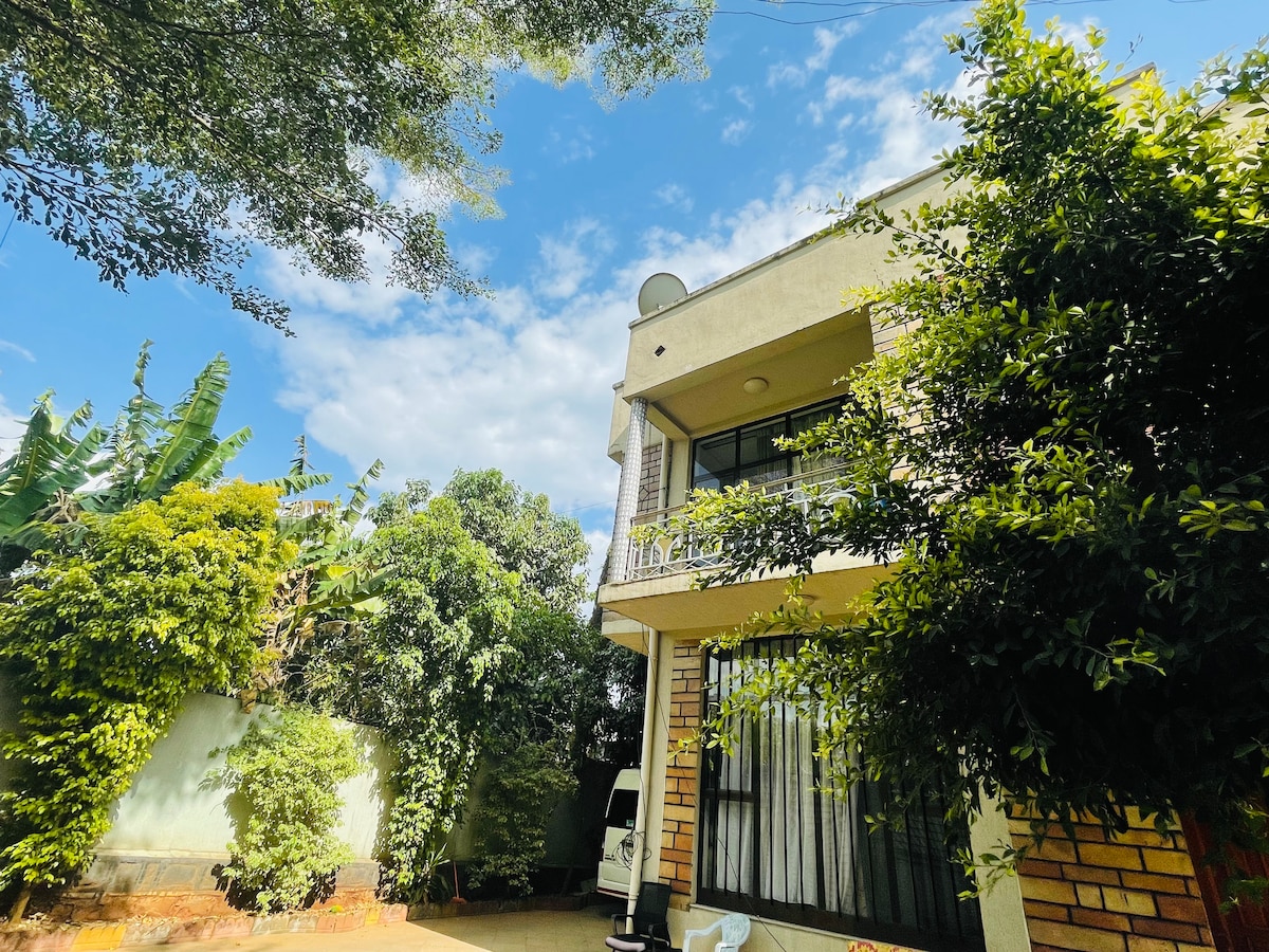A multi-story residence surrounded by lush greenery and trees. The building features a balcony with a railing, offering views of the garden below. The clear blue sky above complements the natural setting, with several plants and foliage enhancing the tranquil environment.