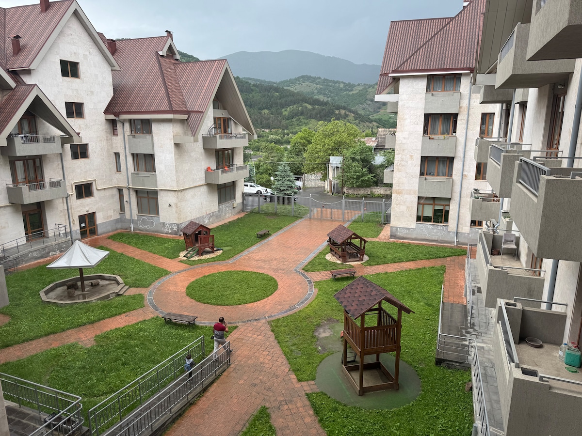 The courtyard is surrounded by multi-story buildings, featuring green grassy areas and brick pathways. Play structures are placed in designated spots, along with seating areas under shaded gazebos. A scenic mountain backdrop is visible in the distance, with trees lining the area.