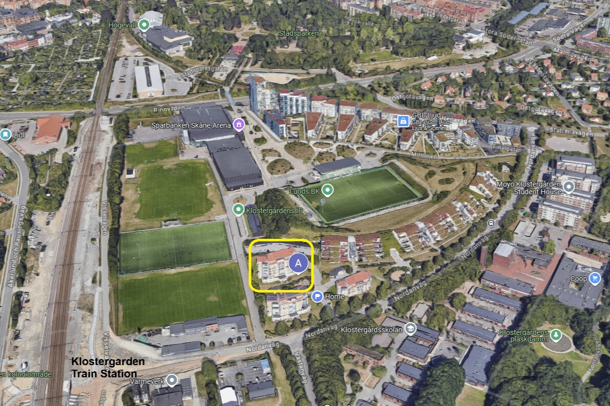 An aerial view captures the surrounding area of Klostergården. The duplex apartment is highlighted, situated amidst green fields and residential buildings, with roads and the nearby Klostergården Train Station visible, indicating the central location within Lund.